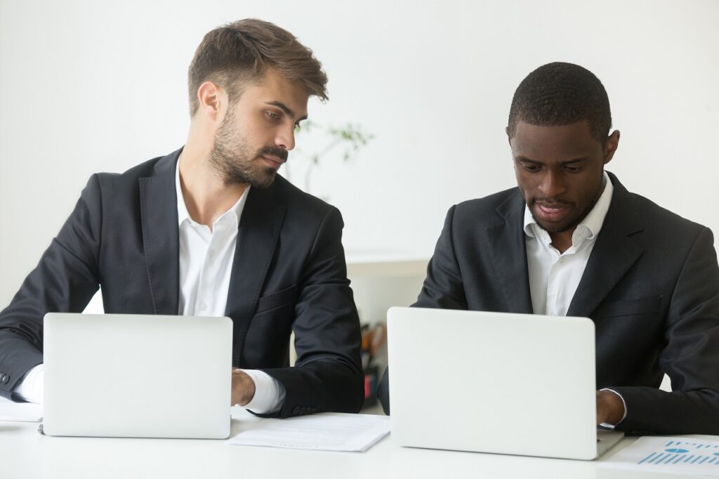 Curious Caucasian male businessman looking at screen of African American colleague busy working at laptop, worker glancing at rival computer, being smart, sly, cunning. Concept of rivalry cooperation