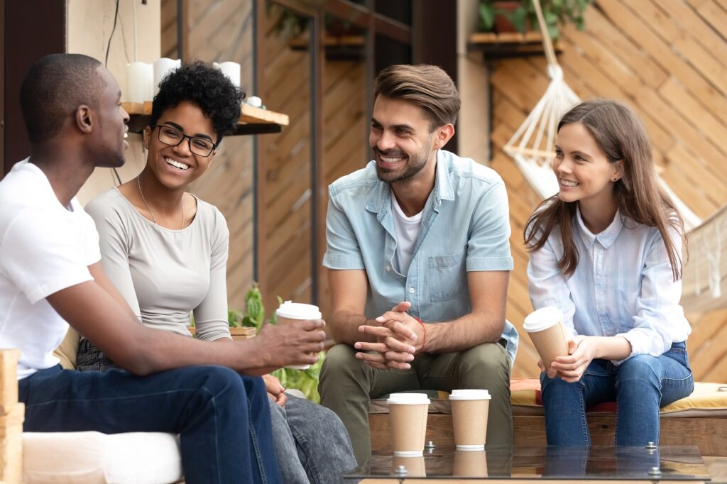Four laughing diverse people positive multi-ethnic friends sitting in cozy cafe summer terrace drink coffee telling funny stories from life feels happy and satisfied enjoy time together on weekend.