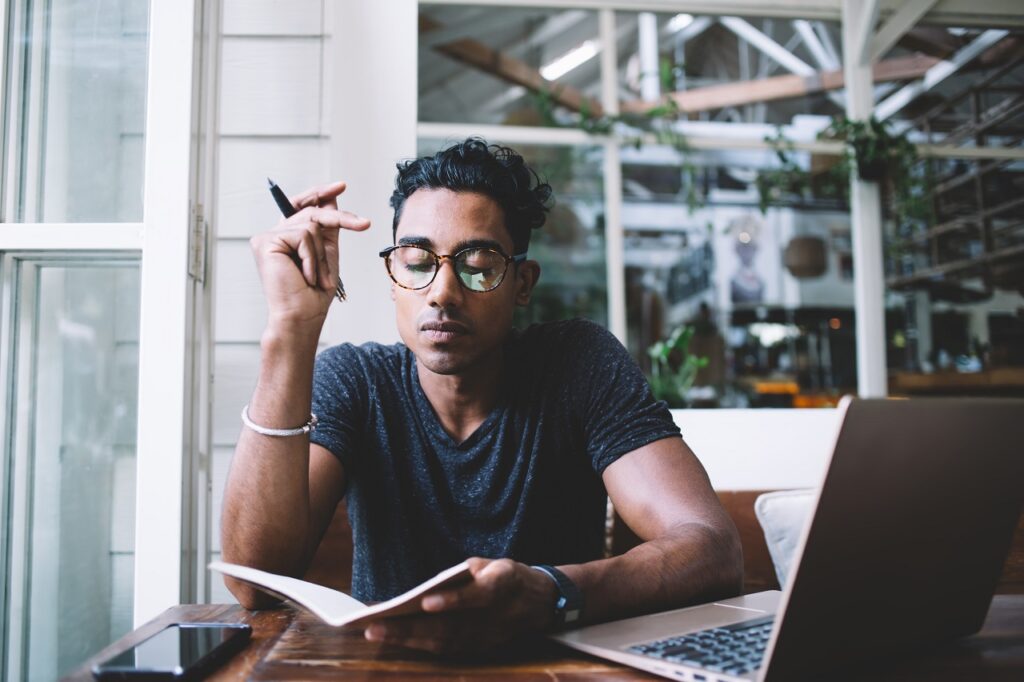 Concentrated serious Hispanic man in eyeglasses thinking and taking notes while sitting at table with laptop and smartphone in tropical cafe