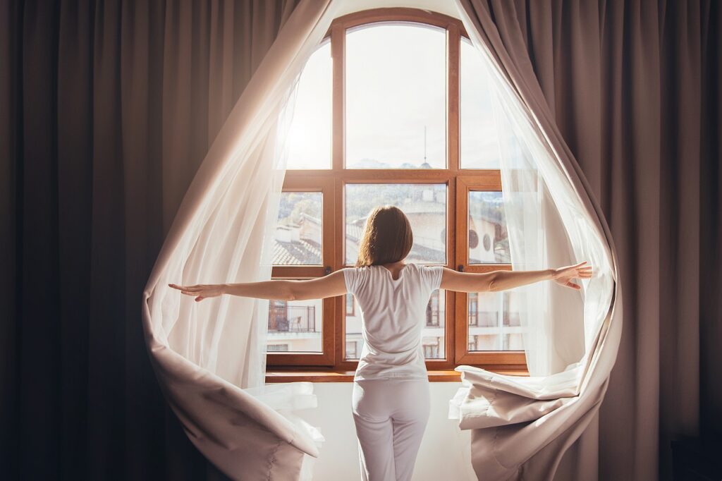 Rear view - young fit woman in home clothes opens curtains of bedroom window. Young girl is ready for new plans and new challenges. Concept of opening yourself to world, new affairs and new promises
