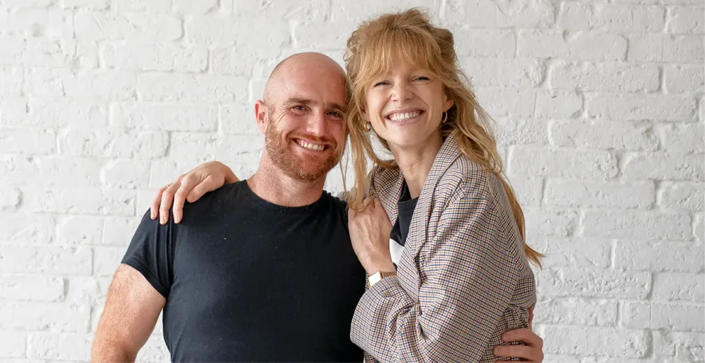 A man and a woman pose together on front of a white brick wall.